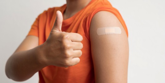 mann mit Orangem T-Shirt und Impfpflaster zeigt Daumen nach oben